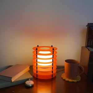 May include: A modern orange table lamp with a stacked disc design. The lamp emits a warm light. A brown mug, a pocket watch, and books are on the wooden surface, creating a cozy atmosphere.