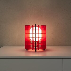 May include: A red, geometric table lamp with a bright, spherical light source. The lamp has a cage-like design with vertical and horizontal bars, and sits on a white surface. The lamp's dimensions are not visible.