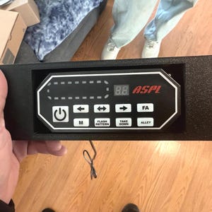 May include: Black control panel with white text and buttons, held by a hand. The panel has the text "ASPL" in red, a digital display, and buttons labeled "M", "Flash Pattern", "Take Down", and "Alley".