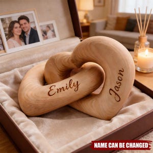 May include: A wooden heart sculpture with the names "Emily" and "Jason" engraved on it. The intertwined hearts are displayed in a brown box with a soft, cream-coloured lining. The text "NAME CAN BE CHANGED" is at the bottom of the box.