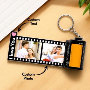 May include: A film roll keychain featuring a black and white film strip design with two rectangular photo slots showing a couple. The strip includes the text I love You and a pink heart. Attached is a black canister labeled Portra 200 EXP 36.