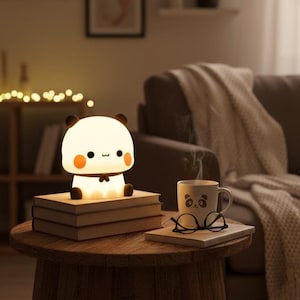 Peut inclure: Une veilleuse en forme de panda est posée sur une pile de livres sur une table en bois. La lumière est blanche avec des oreilles noires et un nœud papillon. Une tasse avec un visage de panda et des lunettes repose sur un livre à proximité.