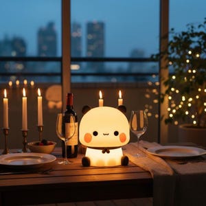 May include: A lit panda-shaped night light sits on a wooden table, surrounded by candles, wine glasses, and a bottle of wine. The panda lamp has black ears, rosy cheeks, and a small bow tie. The background shows a city skyline at dusk.