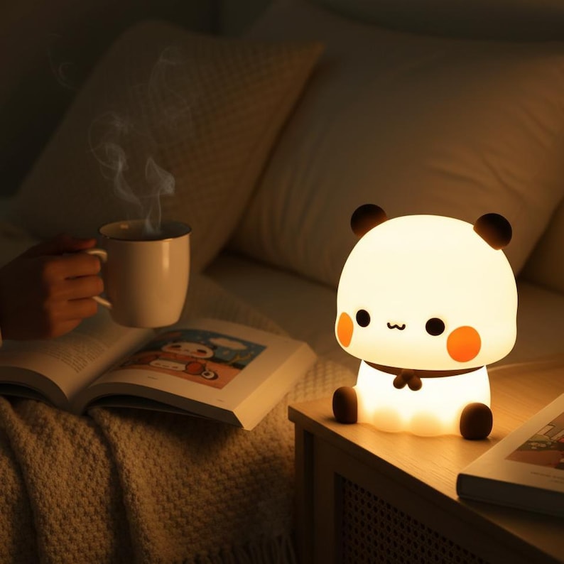 May include: A cute panda-shaped night light with a warm glow sits on a wooden nightstand. The lamp has black ears, eyes, and feet, with orange cheeks. A person holds a white mug with steam rising from it, next to an open book.