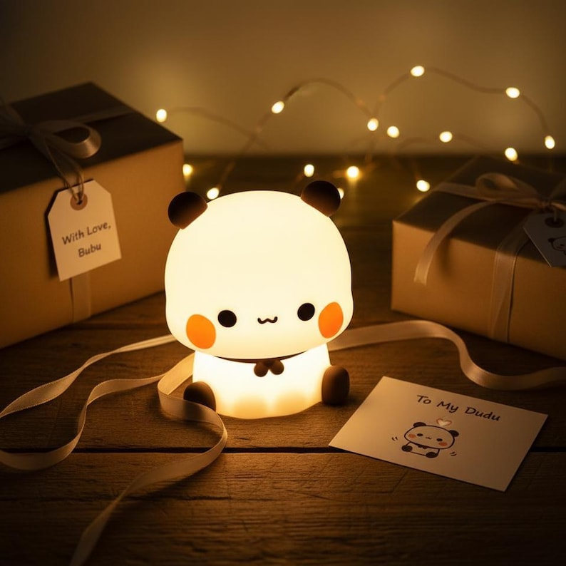 May include: A glowing panda-shaped night light with black ears, eyes, and a bow tie. The panda has orange cheeks and is sitting on a wooden surface. Two wrapped gift boxes and a card that says "To My Dudu" are in the background.