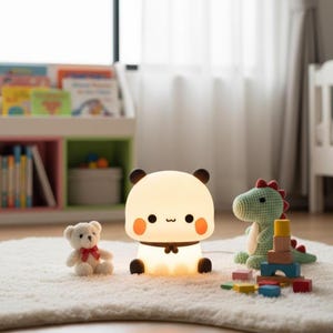 May include: A cute panda-shaped night light with a warm glow, black ears, and orange cheeks. A small teddy bear and a green crocheted dinosaur toy are nearby, along with colorful wooden blocks. The lamp is on a white rug.