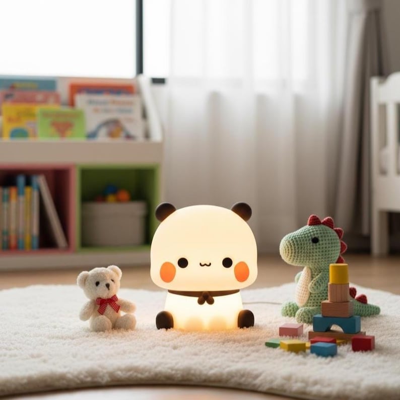 May include: A cute panda-shaped night light with a warm glow, black ears, and orange cheeks. A small teddy bear and a green crocheted dinosaur toy are nearby, along with colorful wooden blocks. The lamp is on a white rug.