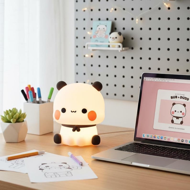 May include: A cute panda-shaped night light with a soft glow sits on a wooden desk. The lamp has black ears, orange cheeks, and a black bow tie. A laptop displays a matching panda graphic. Other items include pens, a succulent, and drawings.