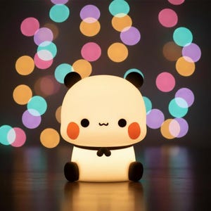 May include: A cute panda-shaped night light with a soft, glowing appearance. The light has black ears, eyes, and feet, with red cheeks. The background features colorful bokeh lights, creating a warm and inviting ambiance. This decorative lamp is perfect for a child's room.