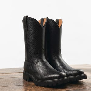 May include: A pair of black leather cowboy boots with a textured pattern. The boots have a tall shaft, a pointed toe, and a chunky sole. The boots are displayed on a wooden surface.
