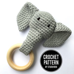 May include: A gray crocheted elephant rattle with a wooden ring. The rattle is attached to the elephant's trunk. The text "CROCHET PATTERN BY STUDIO1859" is on a black circle.