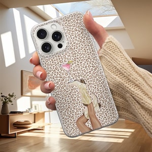 Op de afbeelding: Een telefoonhoesje met een luipaardprint. De hoes heeft een illustratie van een cocktail en cowboylaarzen. De hoes wordt vastgehouden door een persoon die een beige trui draagt.