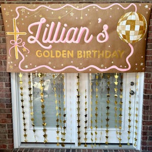Puede incluir: Una pancarta de papel marrón con el texto Lillian's GOLDEN BIRTHDAY en letras rosas y doradas. El diseño incluye una cruz dorada, una bola de discoteca dorada y líneas onduladas rosas con estrellas doradas. Guirnaldas de estrellas cuelgan.