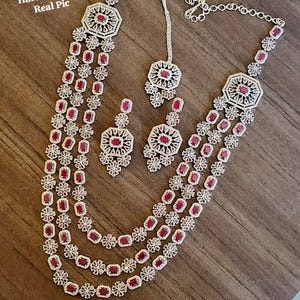 Puede incluir: Un conjunto de collar de tres hilos con pendientes y una pieza para la cabeza a juego. La joyería presenta un metal plateado con piedras rojas rectangulares y cuadradas. El texto en la imagen dice "AD Diamond GJ Polish Heavy Premium Haram - Ruby Real Pic."