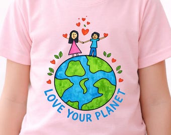 May include: A young girl smiles, wearing a light pink t-shirt. The shirt features a colorful graphic of the Earth with two stick figures holding hands, surrounded by hearts and the words "Love Your Planet."