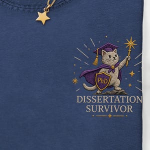Può includere: T-shirt blu navy con un gatto dei cartoni animati con cappello di laurea e mantello, che tiene in mano una bacchetta magica. Il testo "Dissertation Survivor" è sotto. Una collana con pendente a stella dorata è sulla maglietta.