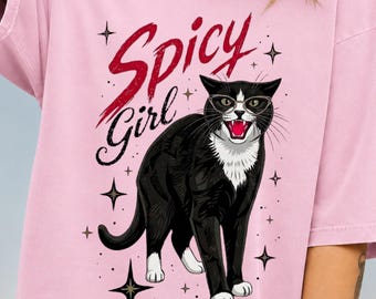 May include: A light pink short-sleeve t-shirt featuring a graphic of a black and white cat wearing glasses with its mouth open. The text "Spicy Girl" is printed in red above the cat, surrounded by small black star icons across the front.