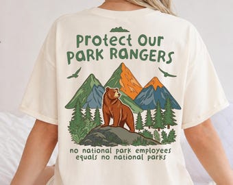 May include: A cream-colored t-shirt featuring a graphic on the back with the text "Protect Our Park Rangers" and "no national park employees equals no national parks." The design includes a brown bear, green pine trees, and colorful mountains.