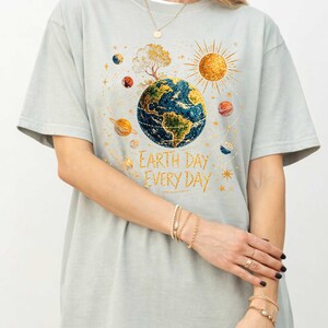 Puede incluir: Camiseta gris claro con un gr&aacute;fico colorido de la Tierra rodeada de planetas y un sol dorado. El texto "Earth Day Every Day" est&aacute; impreso debajo. La camiseta tiene mangas cortas.