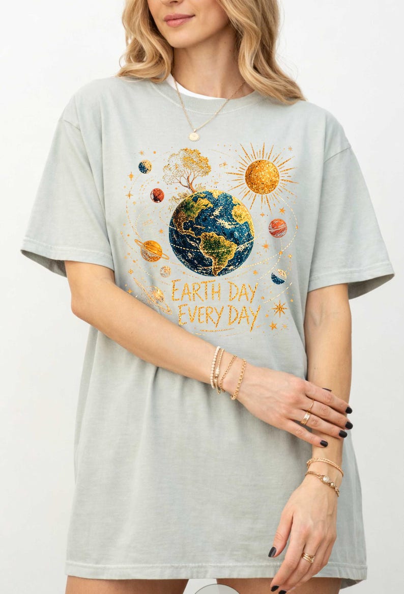 Puede incluir: Camiseta gris claro con un gr&aacute;fico colorido de la Tierra rodeada de planetas y un sol dorado. El texto "Earth Day Every Day" est&aacute; impreso debajo. La camiseta tiene mangas cortas.