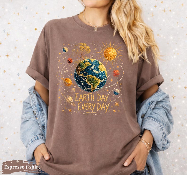 Puede incluir: Camiseta color espresso con un gr&aacute;fico de la Tierra rodeada de planetas y un sol dorado. El texto "Earth Day Every Day" est&aacute; impreso debajo. La camiseta tiene las mangas dobladas.