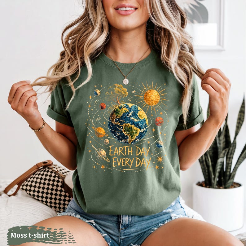 Puede incluir: Camiseta verde musgo con un gr&aacute;fico de la Tierra rodeada de planetas y un sol, con el texto "Earth Day Every Day" en blanco. La camiseta est&aacute; hecha de un material suave.