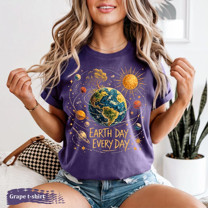 Puede incluir: Una camiseta morada con un gr&aacute;fico de la Tierra rodeada de planetas, un sol y un &aacute;rbol. El texto "Earth Day Every Day" est&aacute; impreso debajo. La camiseta es de color uva.