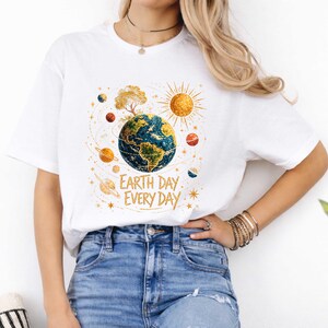 Puede incluir: Camiseta blanca con un gr&aacute;fico colorido de la Tierra, un &aacute;rbol, el sol y planetas. El texto dorado dice "Earth Day Every Day". Esta camiseta informal es adecuada para el uso diario. El dise&ntilde;o promueve la conciencia ambiental.