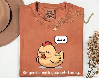 May include: A rust-colored t-shirt featuring a pixelated cartoon chicken with closed eyes and a speech bubble with "Zzz". A gold necklace with a star pendant is on the shirt. The text "Be gentle with yourself today" is at the bottom.
