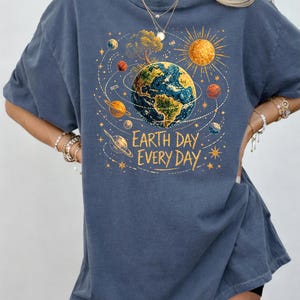 Puede incluir: Camiseta azul con un gr&aacute;fico colorido de la Tierra rodeada de planetas, un sol y un &aacute;rbol. El texto "Earth Day Every Day" est&aacute; impreso debajo. La camiseta tiene mangas cortas y cuello redondo.