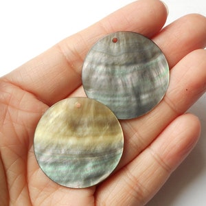 May include: Two round, iridescent, mother-of-pearl shell discs with a hole in the center. The discs are a light brown color with green and black iridescent highlights.