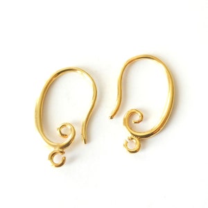 May include: Two gold-colored earring hooks on a white background. Each hook has a curved design with a looped bottom and a decorative swirl at the base of the hook. The hooks are made of a shiny, metallic material.