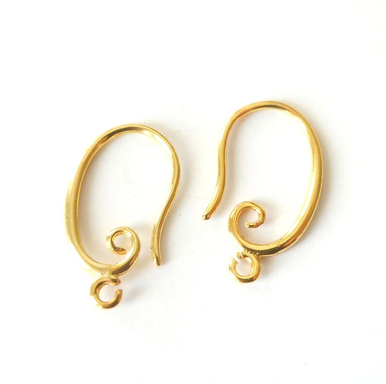 May include: Two gold-colored earring hooks on a white background. Each hook has a curved design with a looped bottom and a decorative swirl at the base of the hook. The hooks are made of a shiny, metallic material.