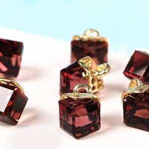 May include: Six small, square, dark red crystal charms with gold accents. The charms have a faceted design and are perfect for jewellery making.