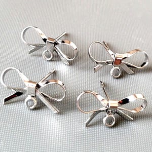 May include: Four silver-colored bow-shaped charms. Each charm has a loop and two small, angled pieces extending from the bow's center. The charms are displayed on a light gray surface.
