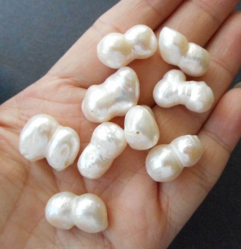 May include: A collection of white baroque pearls, each with an irregular, double-lobed shape. The pearls are arranged on a hand, showcasing their unique and natural beauty.