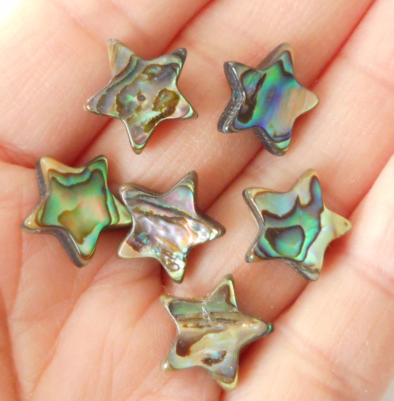 K&ouml;nnte beinhalten: Sechs sternf&ouml;rmige Perlen aus irisierendem Abalone-Muschelschalen. Die Perlen sind in einem Mix aus Gr&uuml;n, Braun und Gold.
