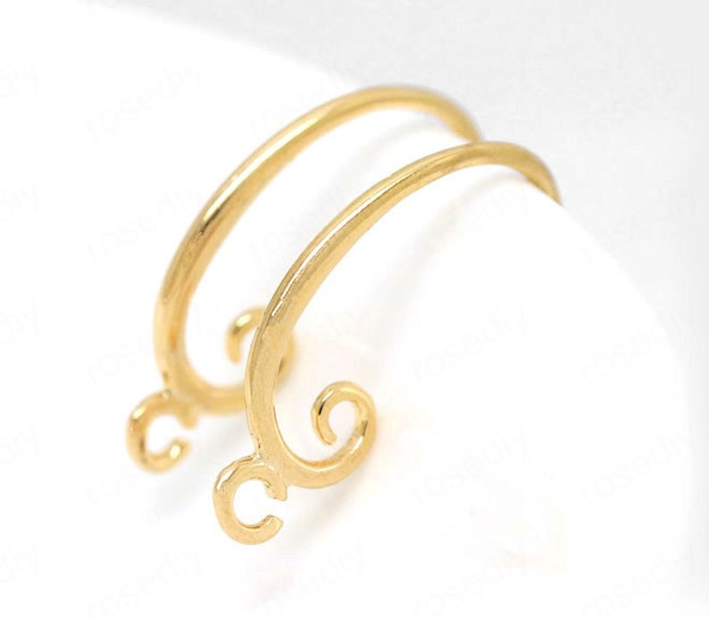 May include: Two gold-colored earring findings with a curved design. Each finding has a loop at the bottom and a decorative swirl at the end of the curved top. The earrings are set against a white background.