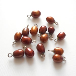 May include: A collection of 14 brown oval-shaped pearls with silver jump rings. The pearls are scattered on a white surface.