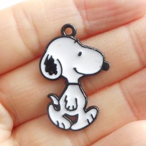 Peut inclure: Breloque en métal noir représentant un chien de dessin animé blanc avec des yeux noirs et un nez noir. Le chien est debout sur ses quatre pattes et porte un collier noir.