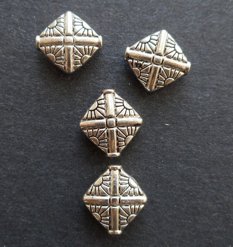 Puede incluir: Cuatro cuentas cuadradas plateadas con un dise&ntilde;o decorativo. Las cuentas tienen una cruz en relieve en el centro y un patr&oacute;n de rayos de sol alrededor de los bordes.