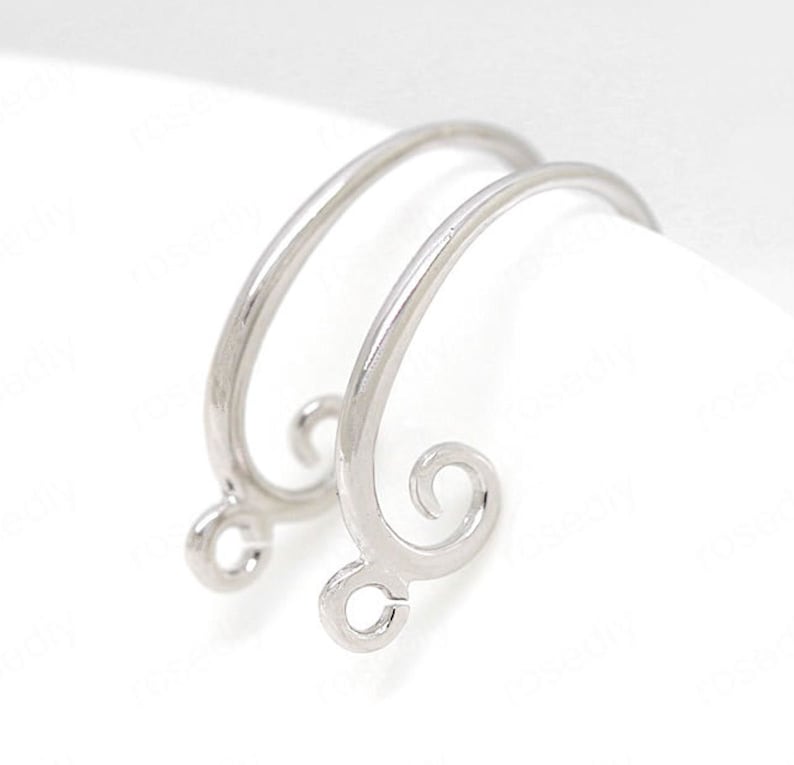 May include: Two silver-colored earring findings. Each finding has a curved, open design with a small loop at the bottom and a decorative swirl at the top. The earrings are set against a white background.
