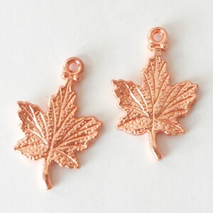 May include: Two copper-coloured maple leaf charms. Each charm features a detailed leaf design with visible veins and a small loop at the top for attaching to jewellery or other crafts. The charms are on a white background.