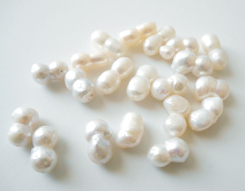 May include: A collection of white baroque pearls, each with an irregular shape and a slightly off-white hue. The pearls are arranged on a white surface.