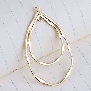 May include: A gold-colored, teardrop-shaped jewelry pendant with a textured, wavy design. The pendant features an inner teardrop shape and a small loop at the top for attaching to a chain or earring hook. The background is a white notebook page.