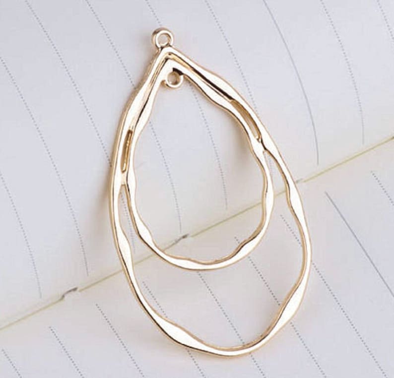 May include: A gold-colored, teardrop-shaped jewelry pendant with a textured, wavy design. The pendant features an inner teardrop shape and a small loop at the top for attaching to a chain or earring hook. The background is a white notebook page.