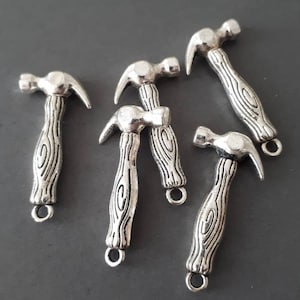 May include: Five silver-toned hammer charms with a wood-grain design on the handle. Each charm has a jump ring for attaching to a necklace, bracelet, or other jewelry.