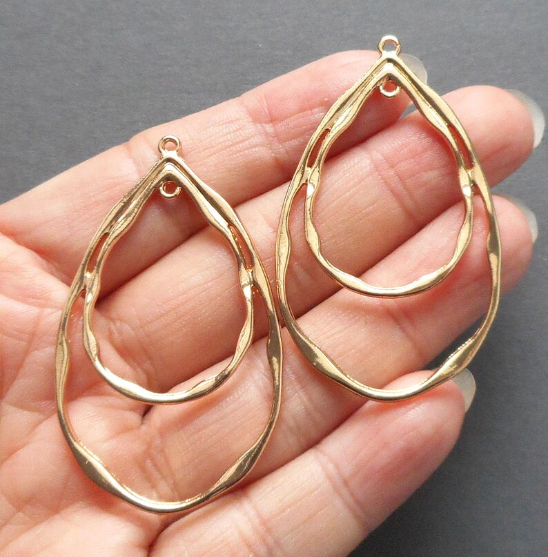 May include: Two gold-colored, open-frame teardrop-shaped earrings. Each earring features a double-layered design with a textured, wavy surface. The earrings have a small loop at the top for attachment.