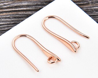 May include: Two rose gold earring hooks. Each hook has a curved design with a small loop at the bottom for attaching charms or beads. The hooks are displayed against a white and wood grain background.
