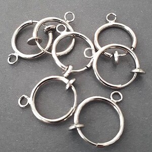 May include: A collection of silver-colored hoop earring findings. Each earring finding is a small, circular hoop with a hinged clasp and a small loop at the top for attaching to other jewelry components. The earrings are displayed on a gray surface.